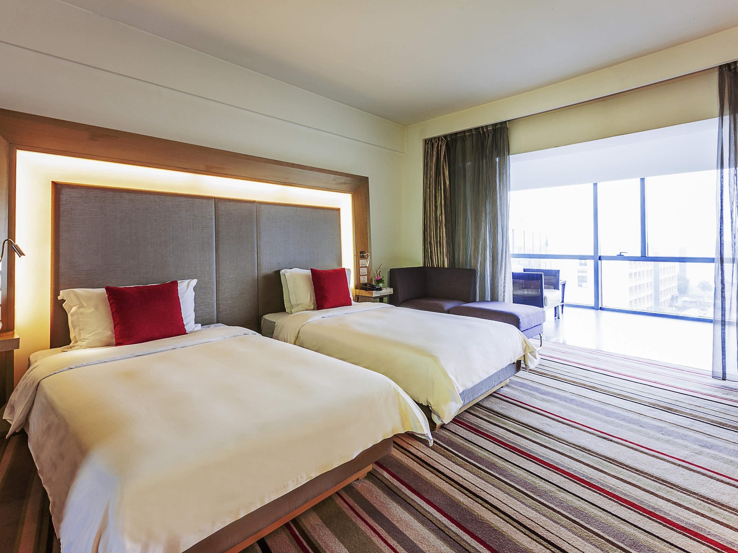 A hotel room with two beds, a large window with curtains, a sofa, and a view of the city.
