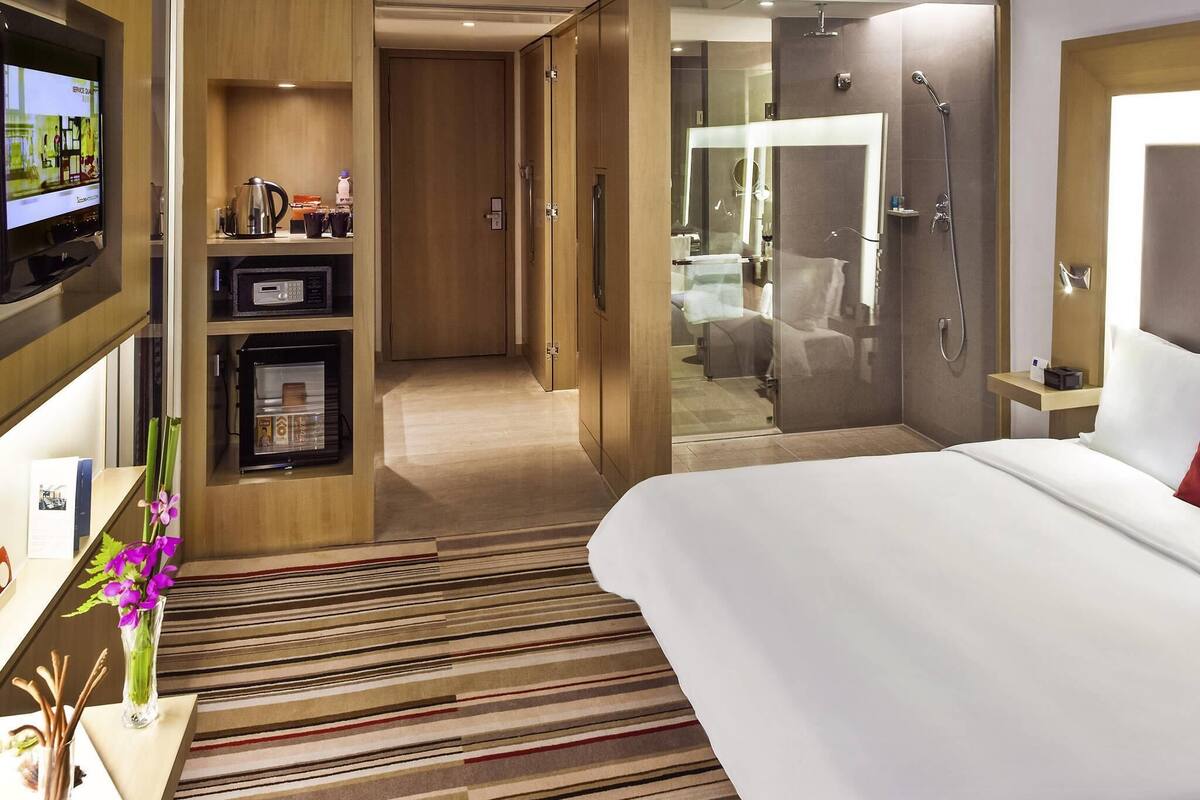 Une chambre d’hôtel comprenant un lit, une télévision, un minibar, une salle de bain avec douche et un couloir menant à une autre chambre.
