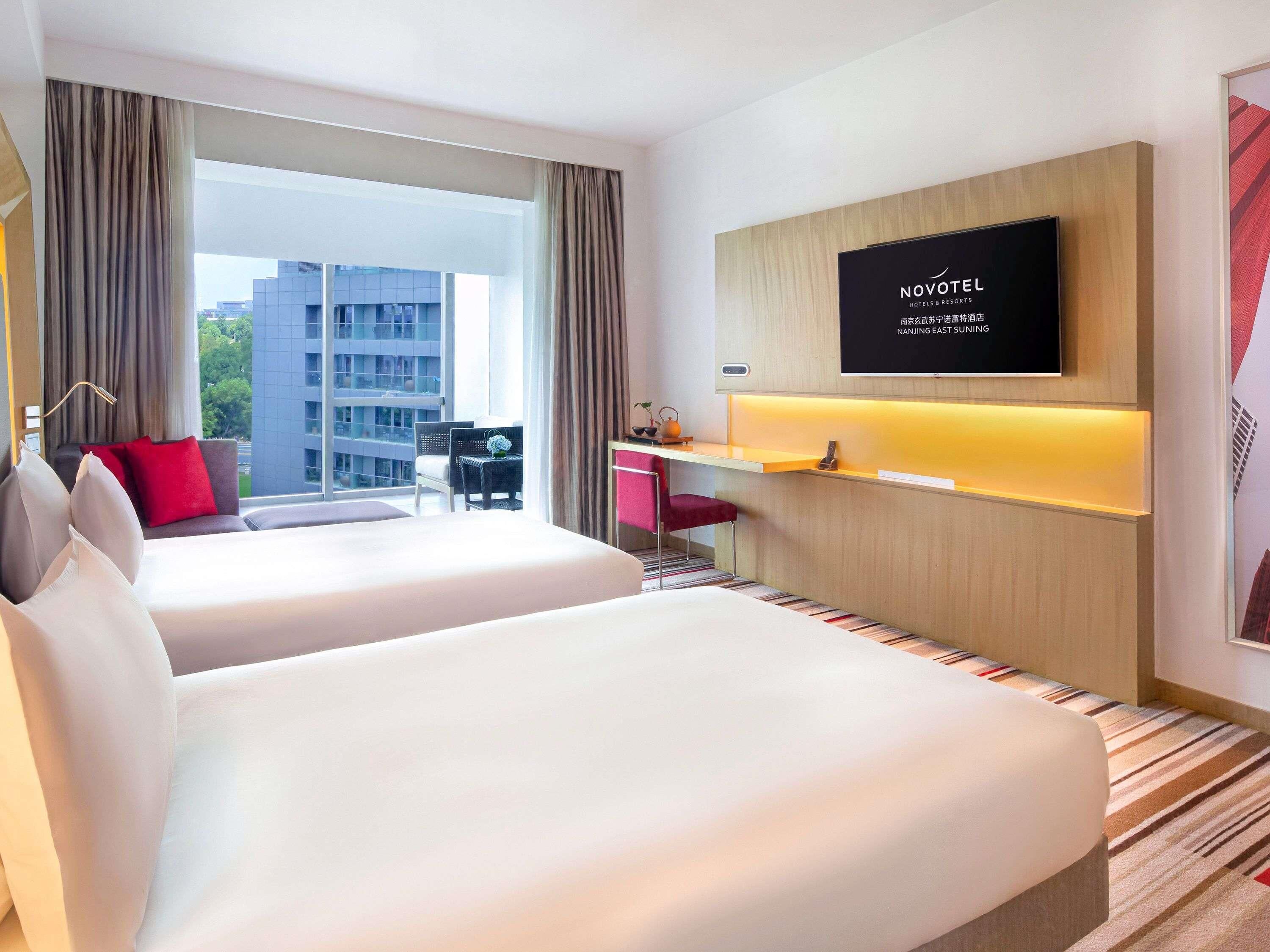 Ein Hotelzimmer mit zwei Betten, einem Schreibtisch, einem Fernseher und Blick auf ein Gebäude.