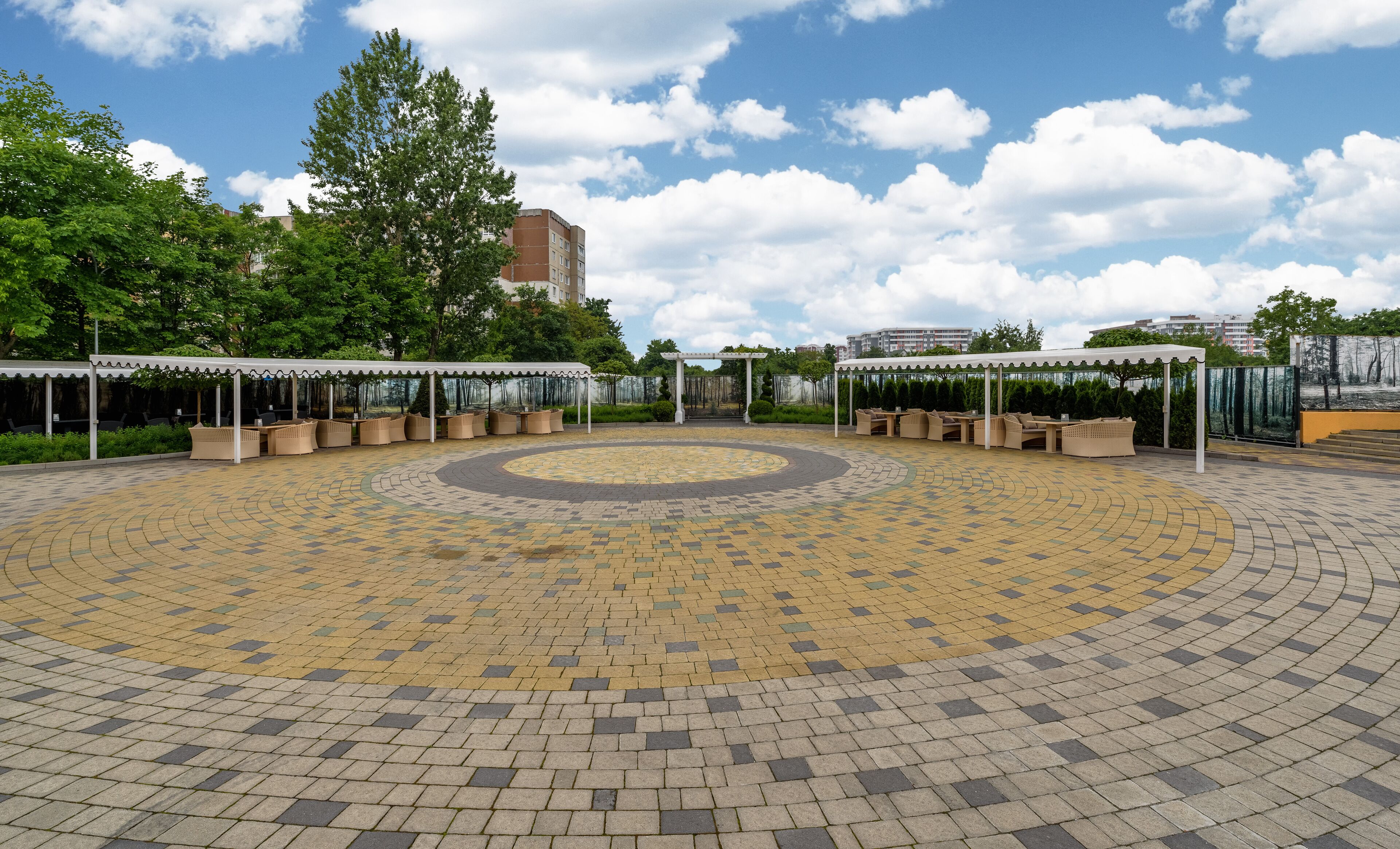 A circular outdoor seating area with a patterned floor, surrounded by covered seating and greenery.