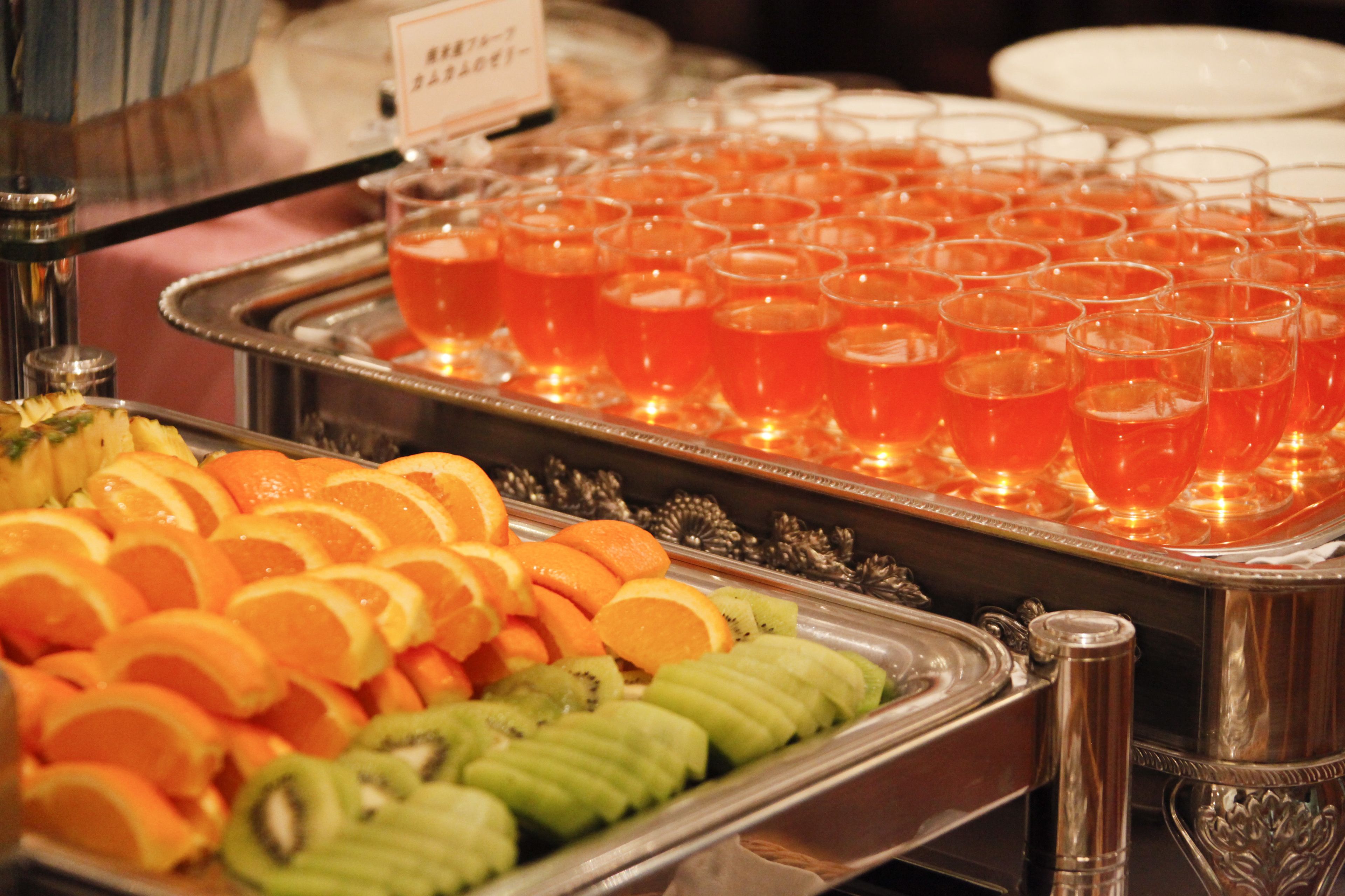 A buffet with sliced oranges, kiwi, and a tray of red punch glasses.