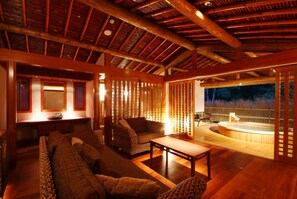 Een knus interieur met een houten plafond, een zithoek met banken en een tafel, en een zichtbare zone met een jacuzzi, afgescheiden door een scheidingswand.