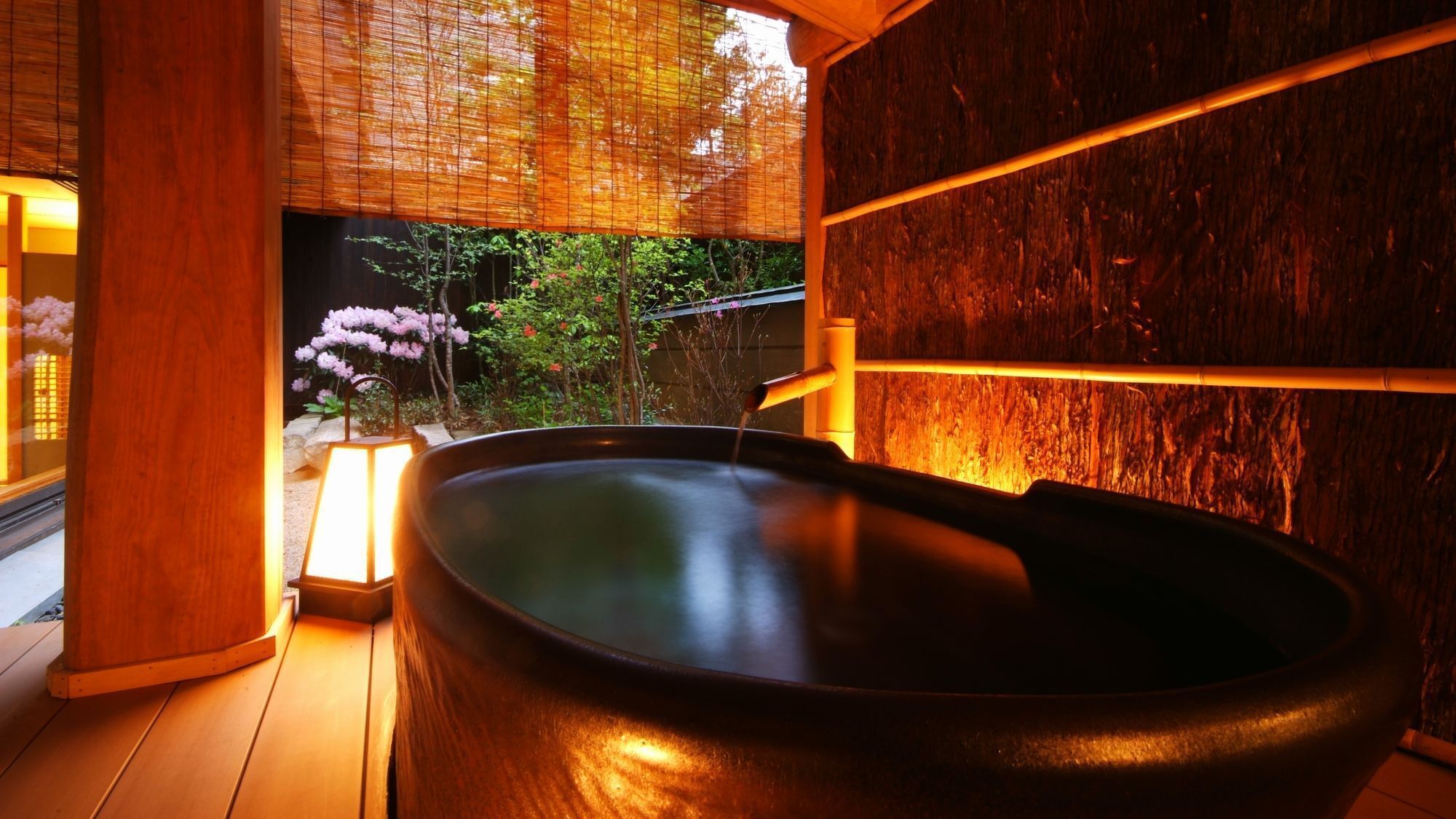 Un bain à remous avec un intérieur en bois, un paravent en bambou et une vue sur un jardin aux fleurs roses.