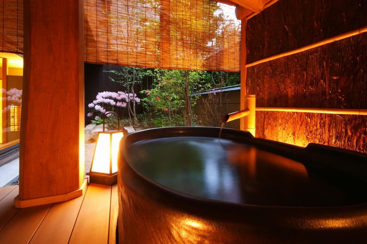 Een hottub met een houten binnenkant, een bamboeblind en uitzicht op een tuin met roze bloemen.