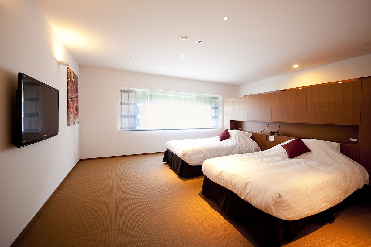 Hotelkamer met twee bedden, een flatscreen televisie aan de muur, een raam met zonwering en een houten hoofdbord.