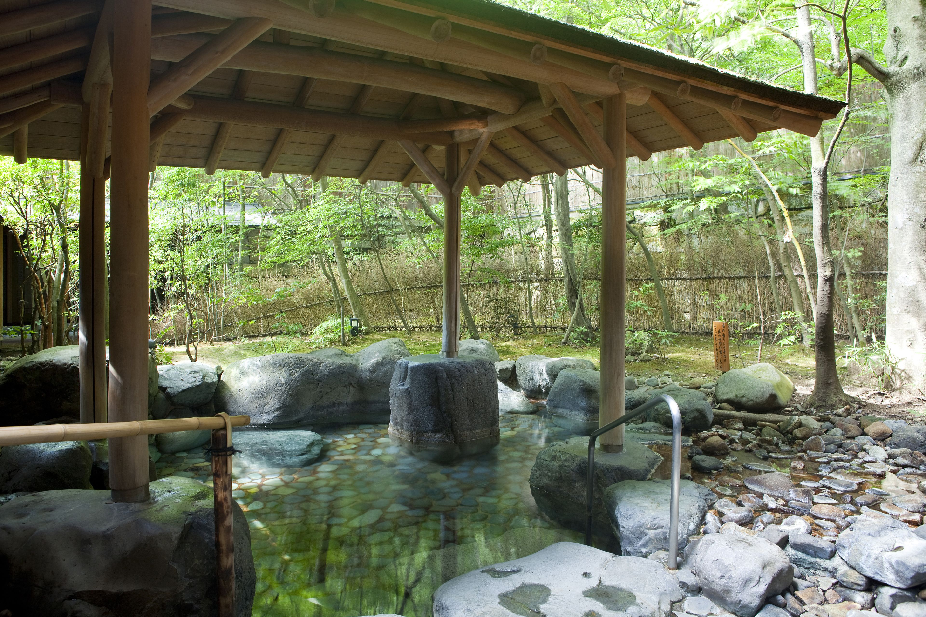 A traditional Japanese onsen with a wooden pavilion, a stone hot spring, and a rocky landscape.