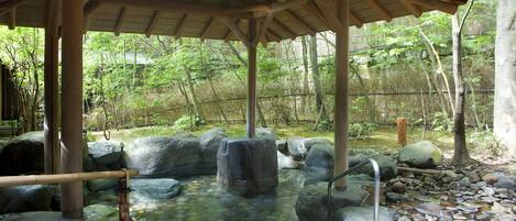 Ein traditionelles japanisches Onsen mit einem hölzernen Pavillon, einer Stein-Thermalquelle und einer felsigen Landschaft.