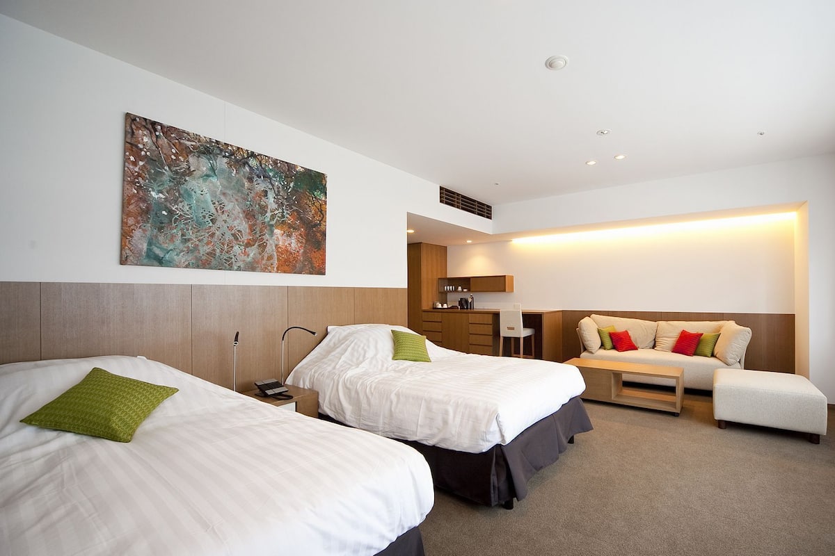 Een hotelkamer met twee bedden, een bureau, een stoel, een kleine kitchenette en een groot raam.