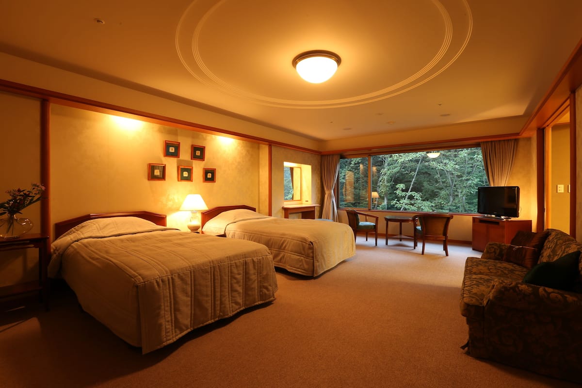 Een hotelkamer met twee bedden, een bureau, een televisie en een groot raam met uitzicht op bomen.