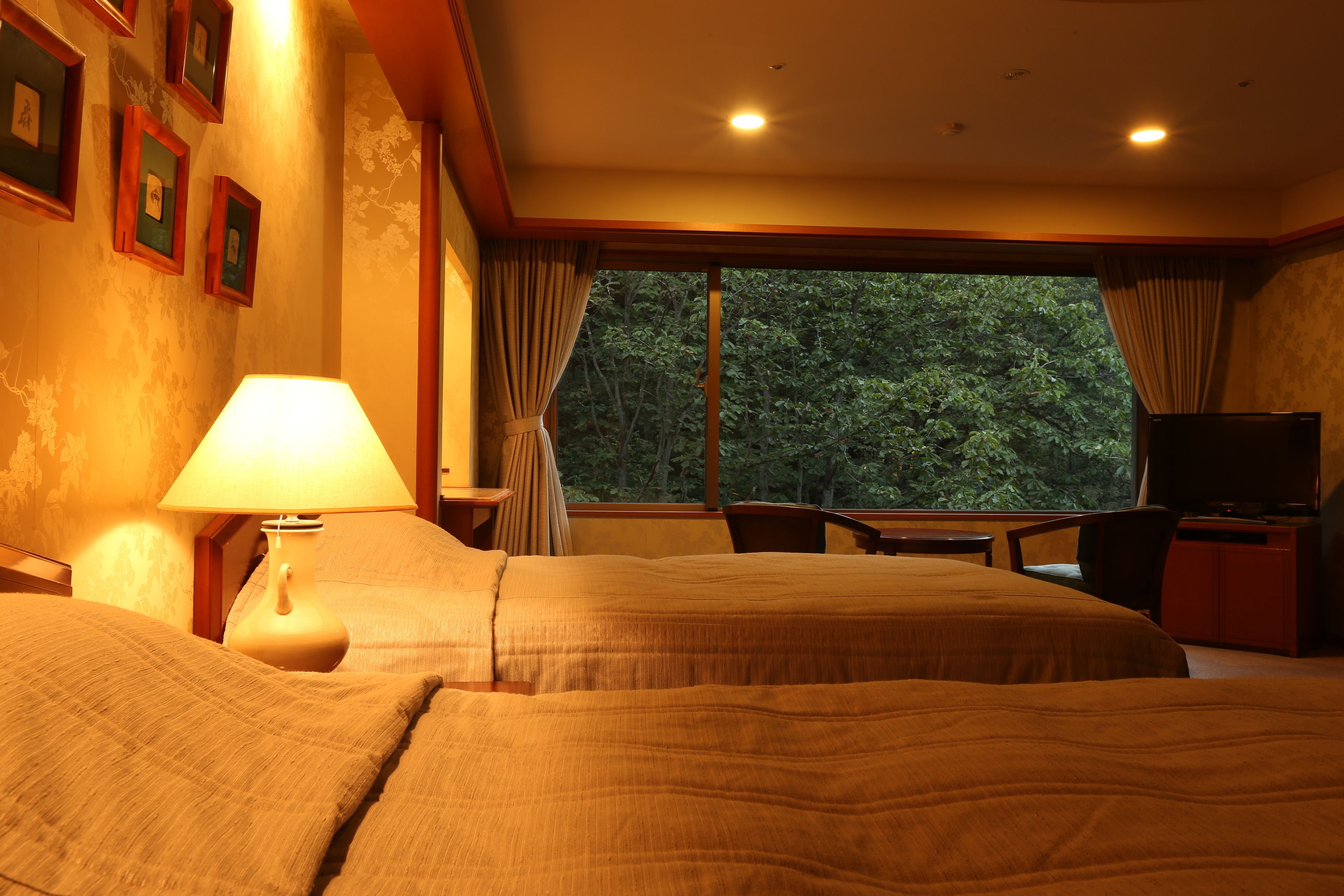 A hotel room with two beds, a desk, a TV, and a window with a forest view.