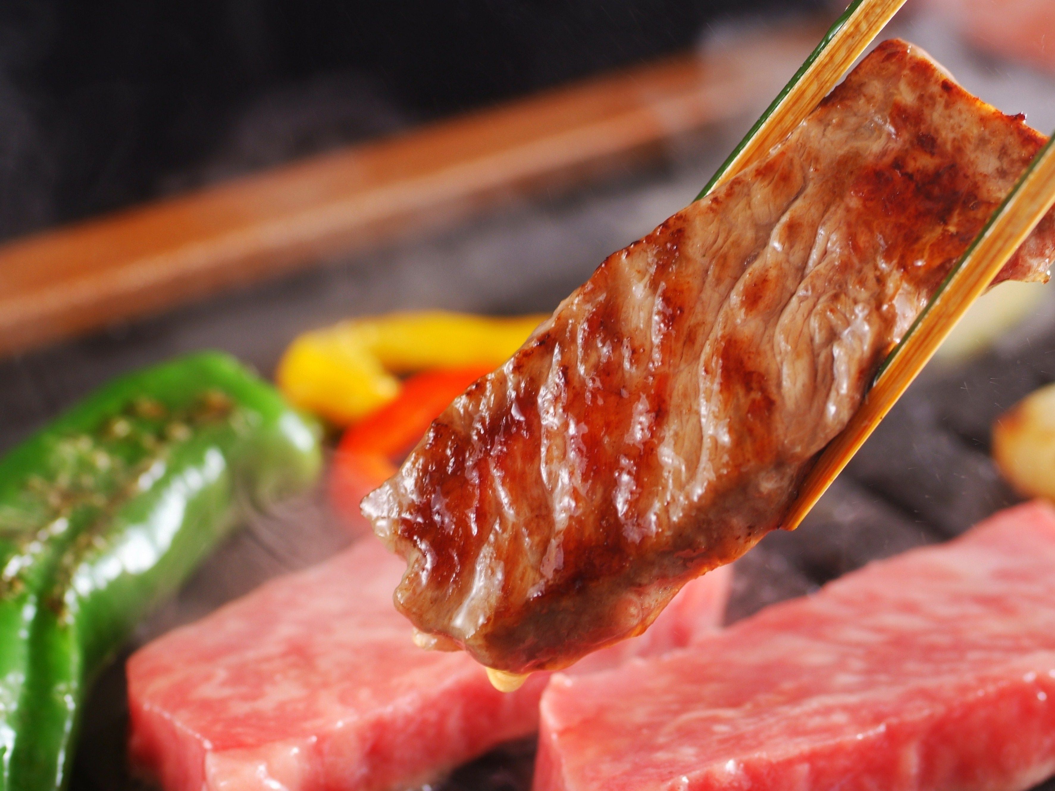 Gros plan sur de la viande grillée tenue à l’aide de baguettes, avec des légumes visibles en arrière-plan.