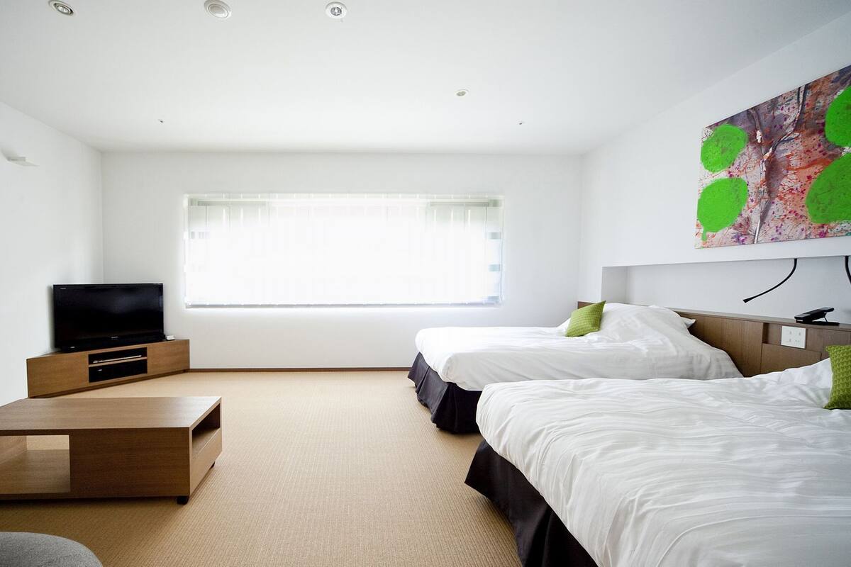 Een moderne hotelkamer met een flatscreen-televisie, een houten bijzettafeltje, twee bedden en een groot raam met luxaflex.