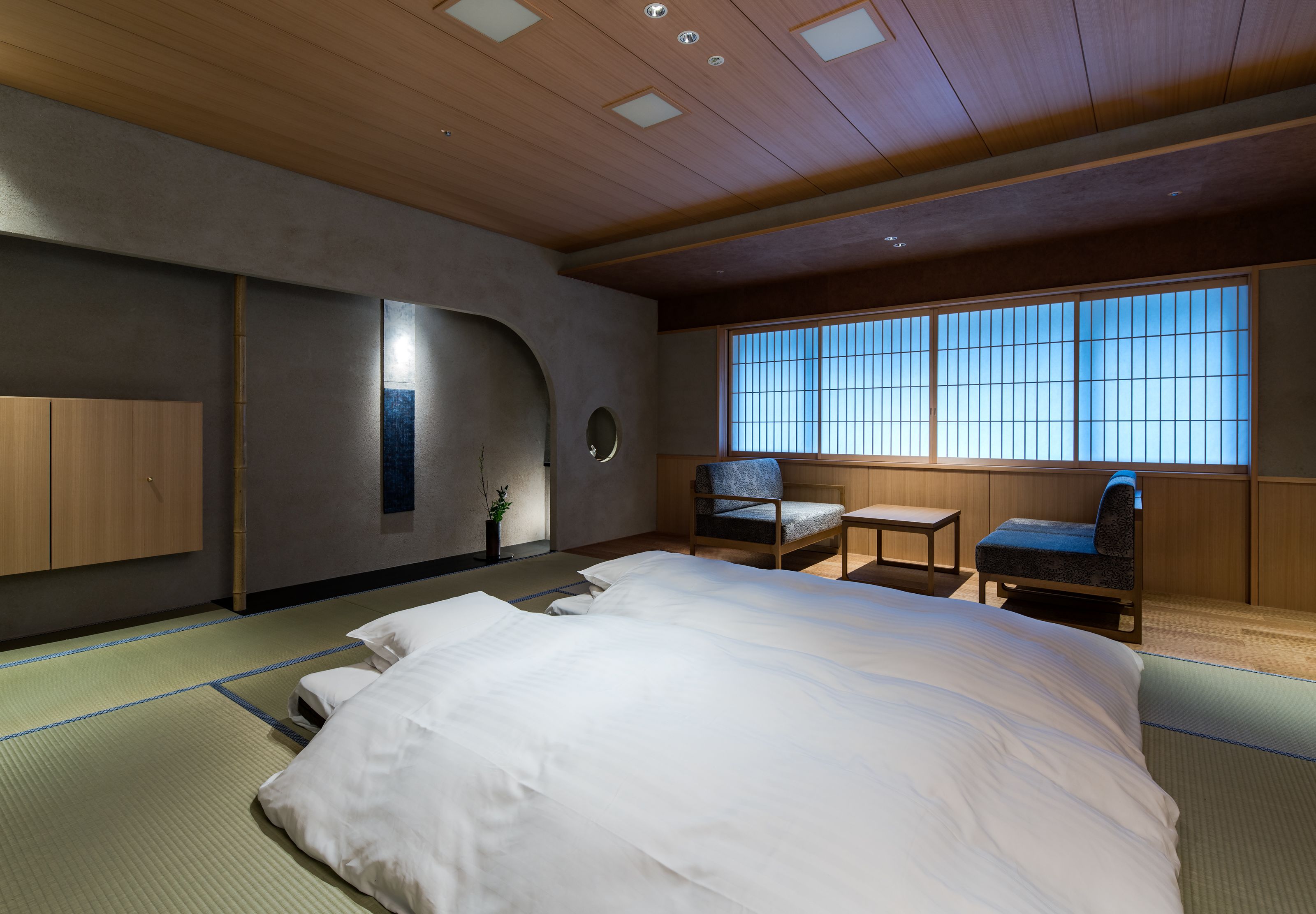 A traditional Japanese-style room with tatami flooring, a large bed, a small table, and a chair.