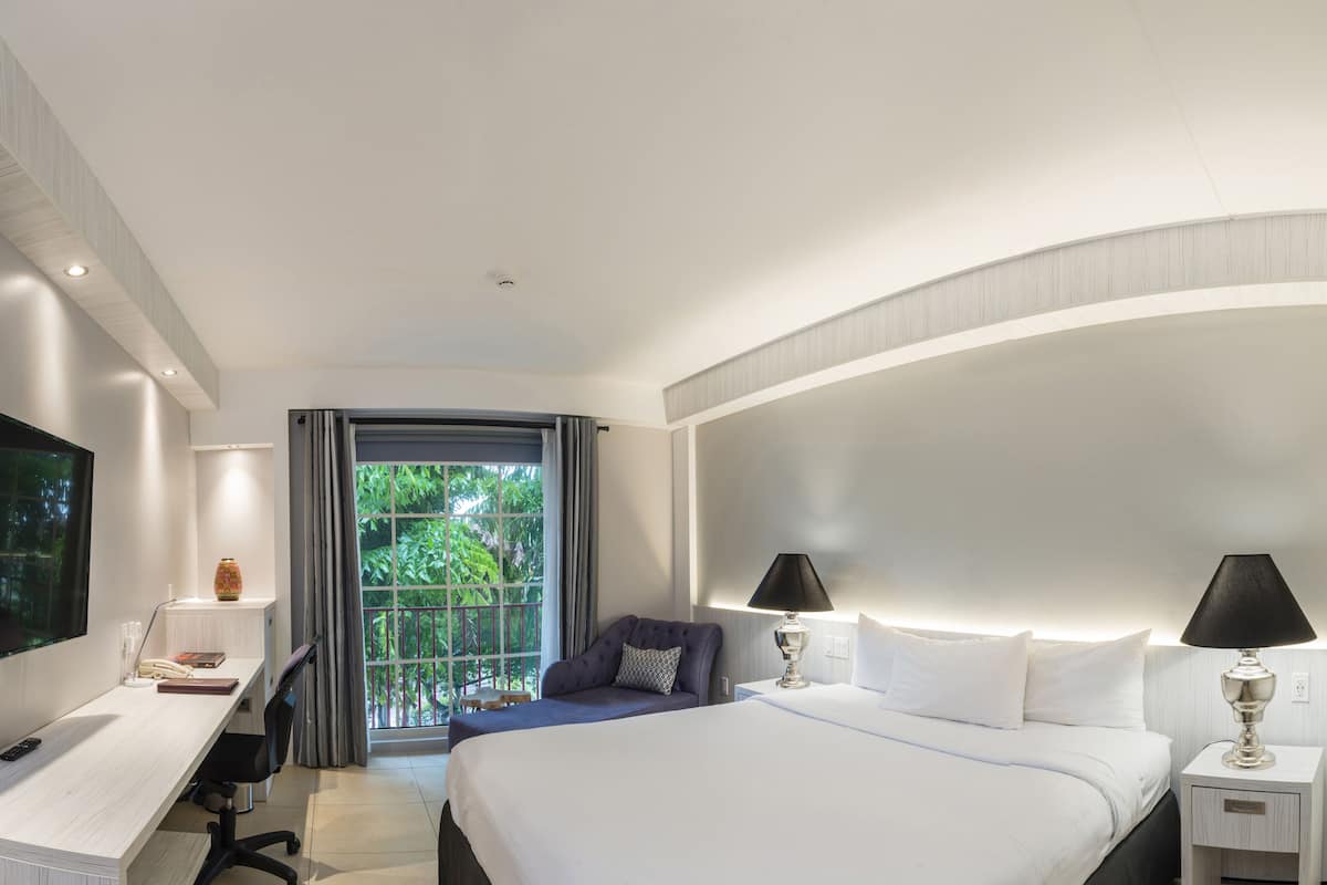 A modern hotel room with a large bed, a desk with a TV, and a balcony with a view of greenery.