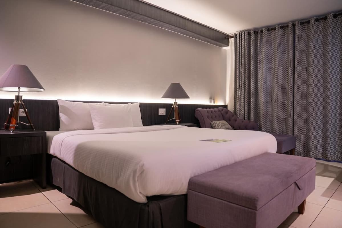 A modern hotel room with a large bed, two bedside lamps, a nightstand, a chair, and a window with patterned curtains.