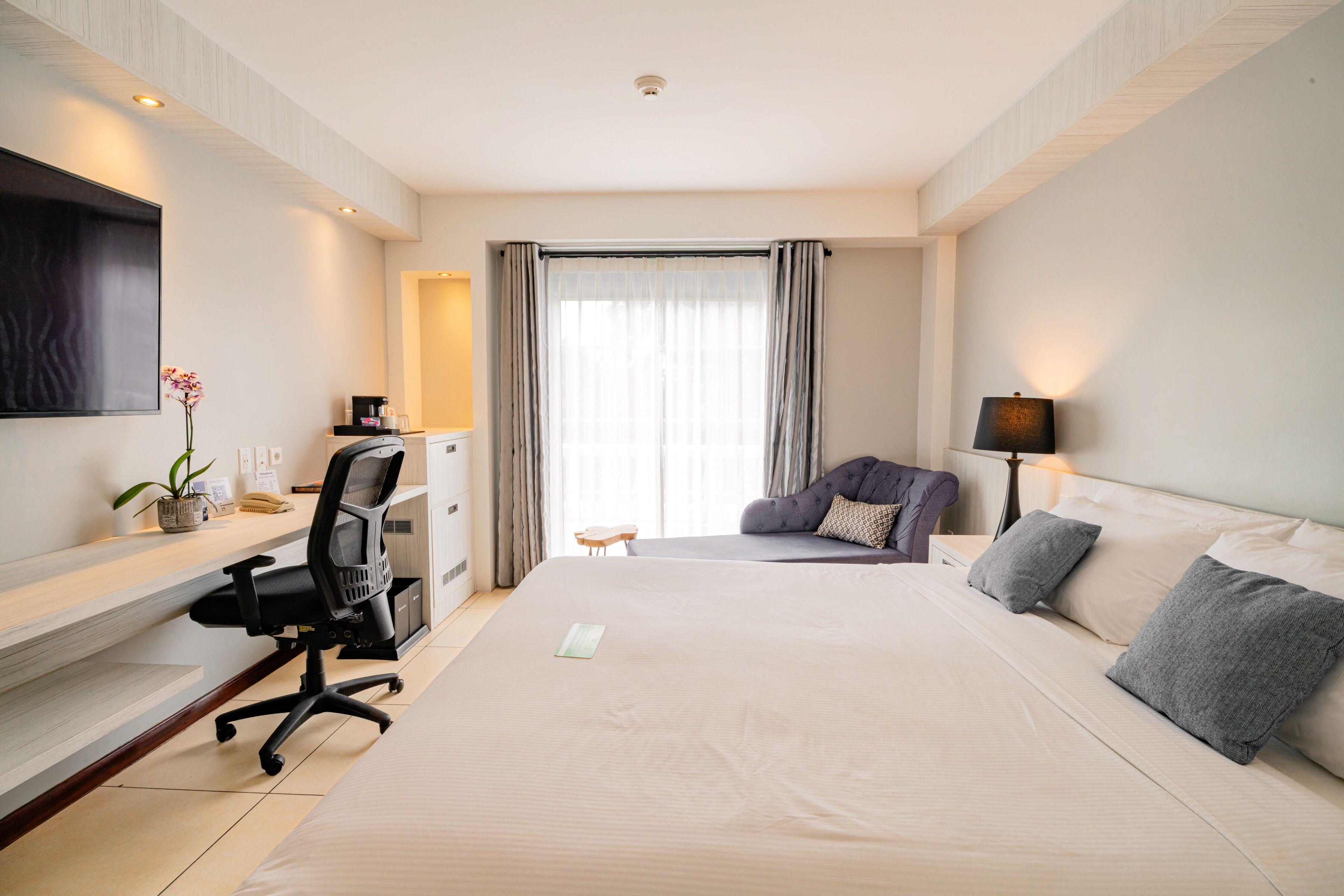 Quarto de hotel com uma cama grande, uma mesa com cadeira, televisão, luminária e janela com cortinas.