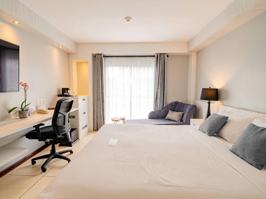 Quarto de hotel com uma cama grande, uma mesa com cadeira, televisão, luminária e janela com cortinas.