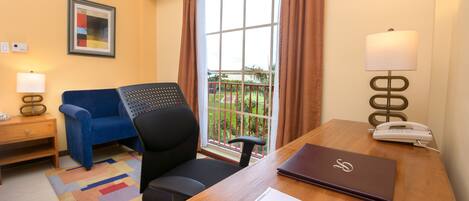 A hotel room with a wooden desk, a blue armchair, a laptop, a telephone, and a window with curtains.