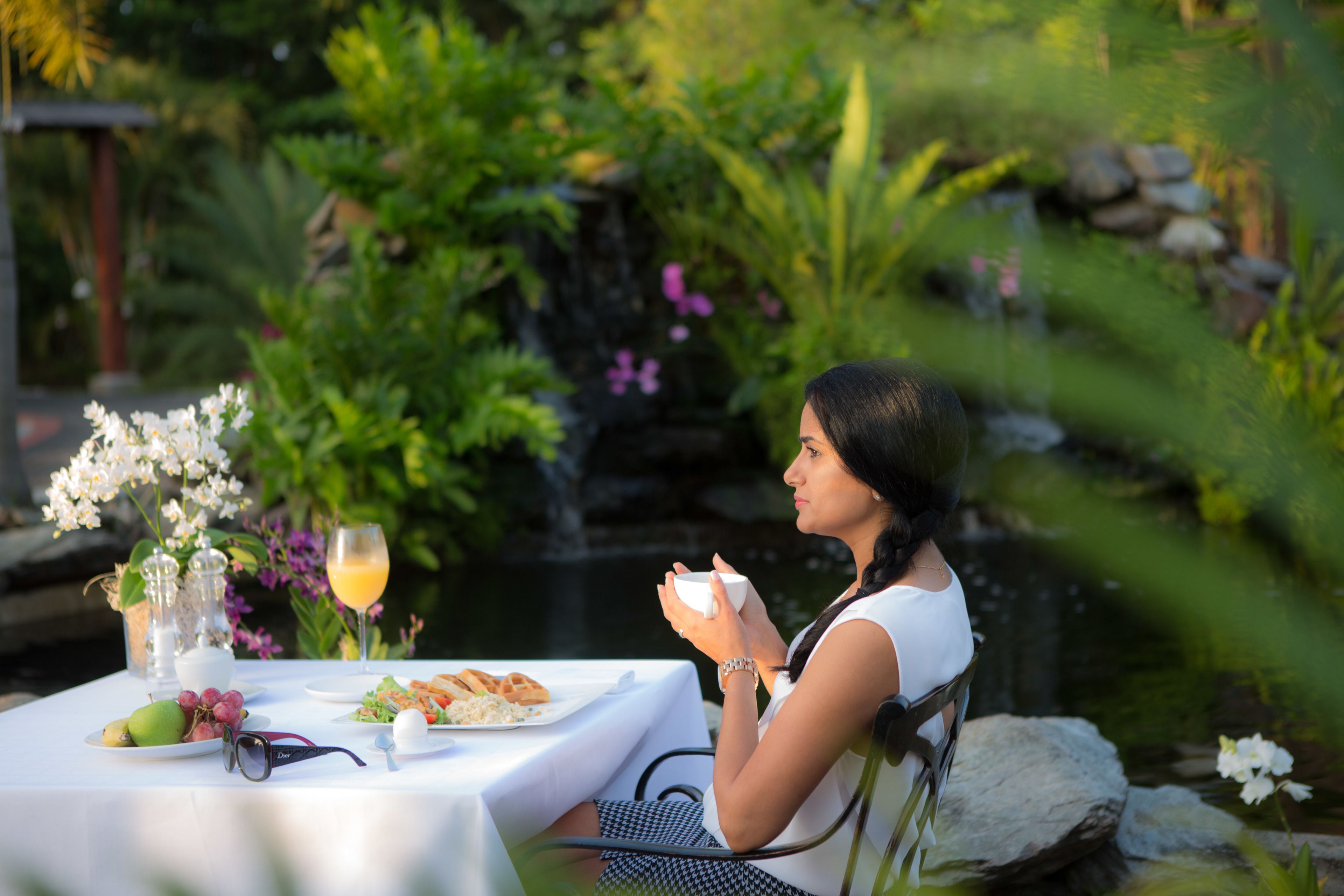 Una mujer sentada en una mesa al aire libre con comida, rodeada de vegetación y un estanque.