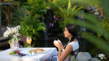 Una mujer sentada en una mesa al aire libre con comida, rodeada de vegetación y un estanque.