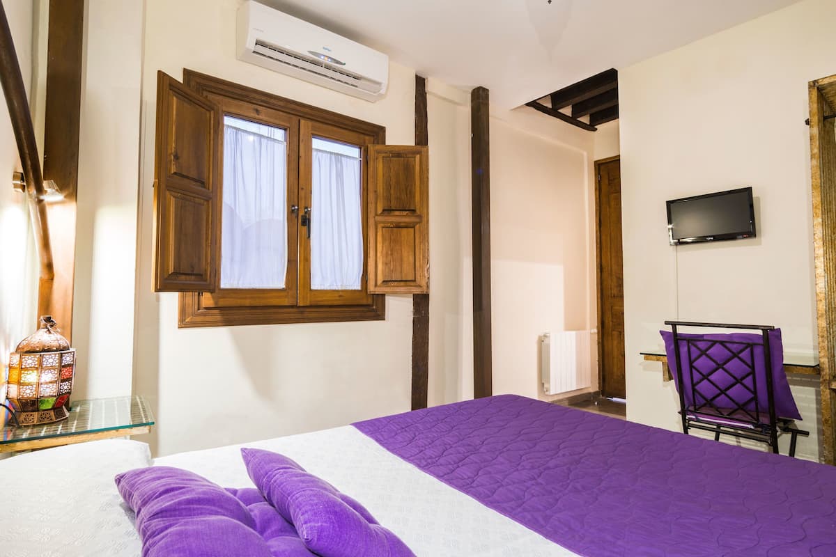 Un dormitorio con cama, ventana con contraventanas de madera, televisor en la pared y una silla.