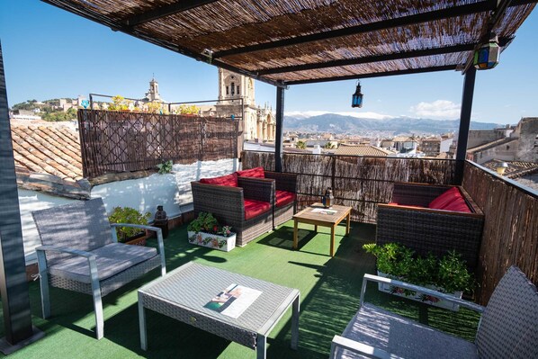 A rooftop terrace with wicker furniture, green artificial turf, and a view of a cityscape and mountains.