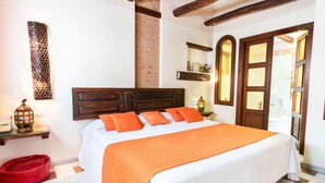 Una cama cuidadosamente tendida con colcha y almohadas naranja, un cabecero de madera, una cama para mascotas roja, una lámpara fijada a la pared y una puerta con paneles de vidrio.
