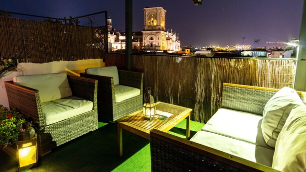 Terraza en la azotea con mobiliario de mimbre, una mesa con un farol y vistas a un paisaje urbano nocturno.