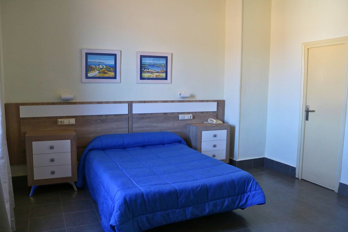 A bedroom with a blue bedspread, two framed pictures on the wall, a bedside table with a phone, and a door to the right.