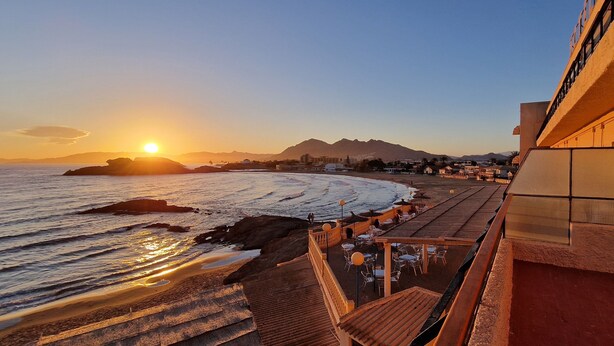 Vista costera con atardecer, una playa y un edificio con balcón que da al océano.