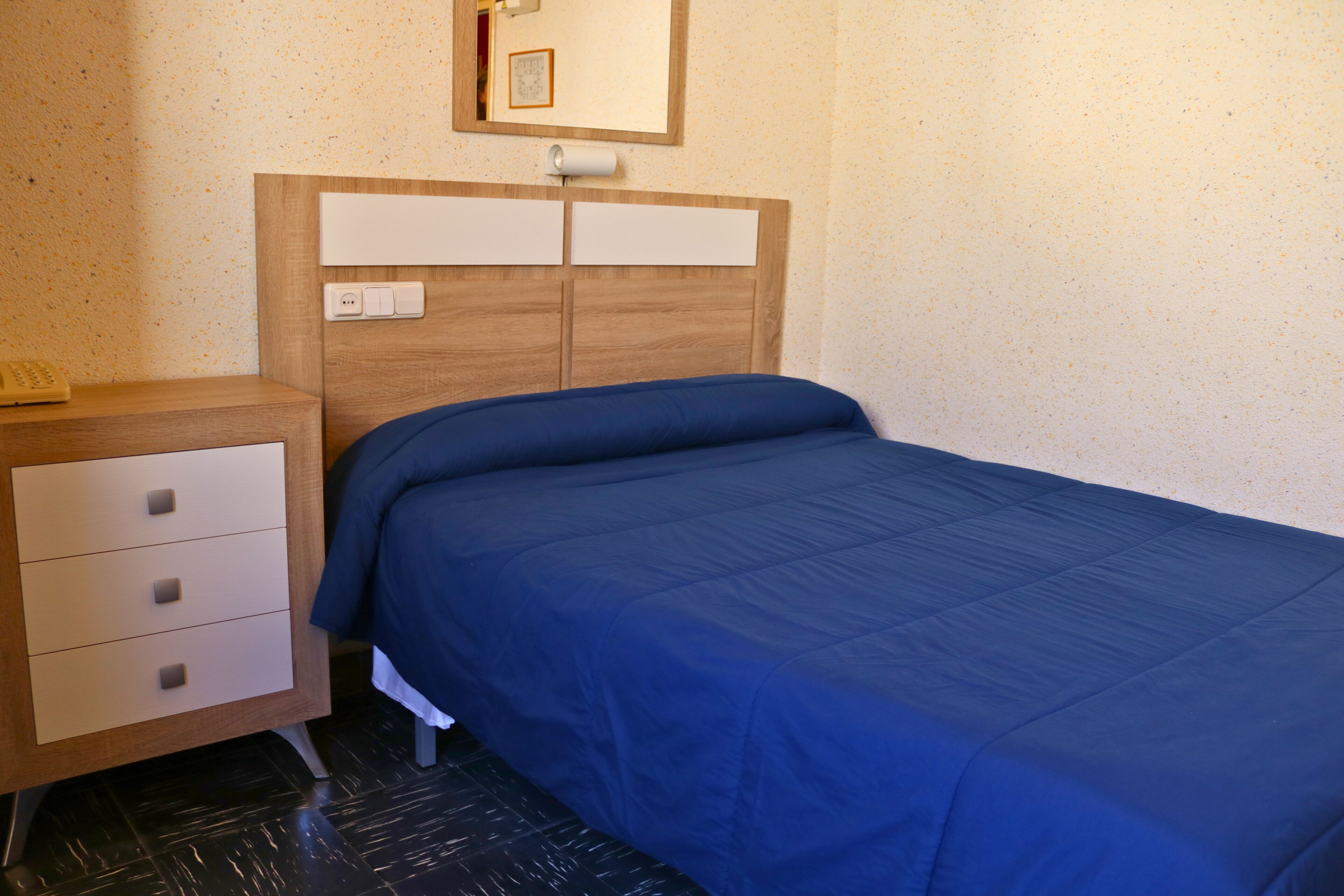 A bedroom with a wooden headboard, a bed with a blue bedspread, a bedside table with a lamp, and a wall-mounted light switch.