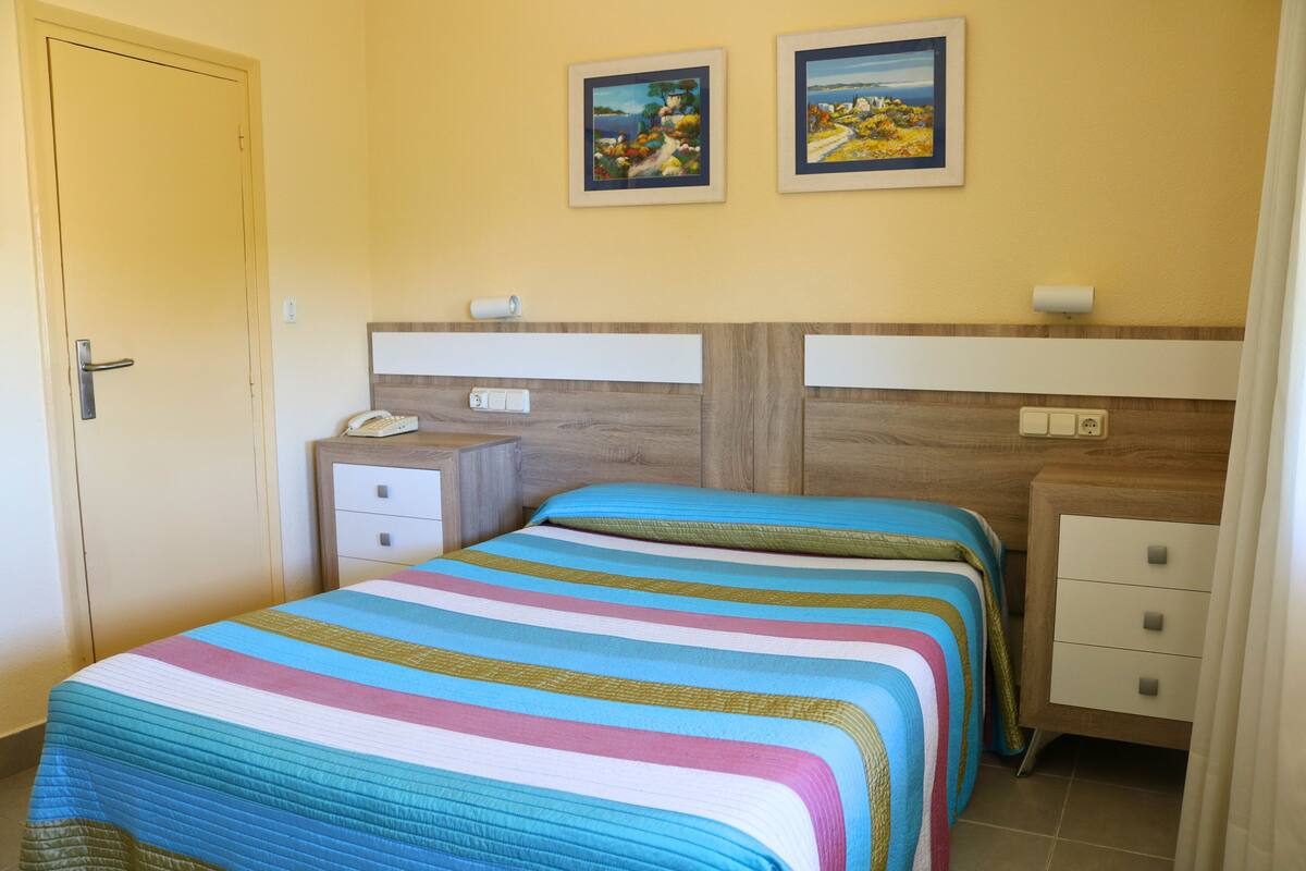 A bedroom with a bed, two bedside tables, a nightstand, a door, and two framed pictures on the wall.