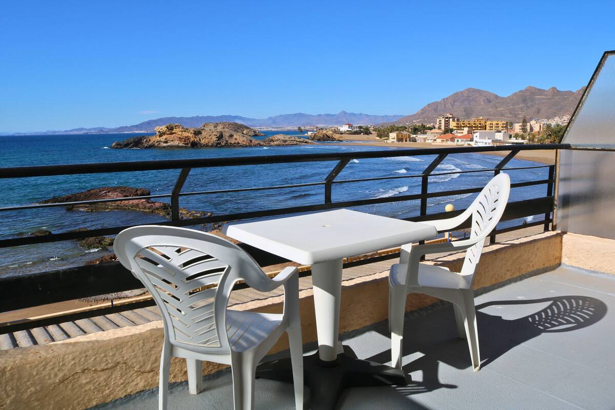 A balcony with a table and two chairs overlooking a coastal area with mountains in the background.