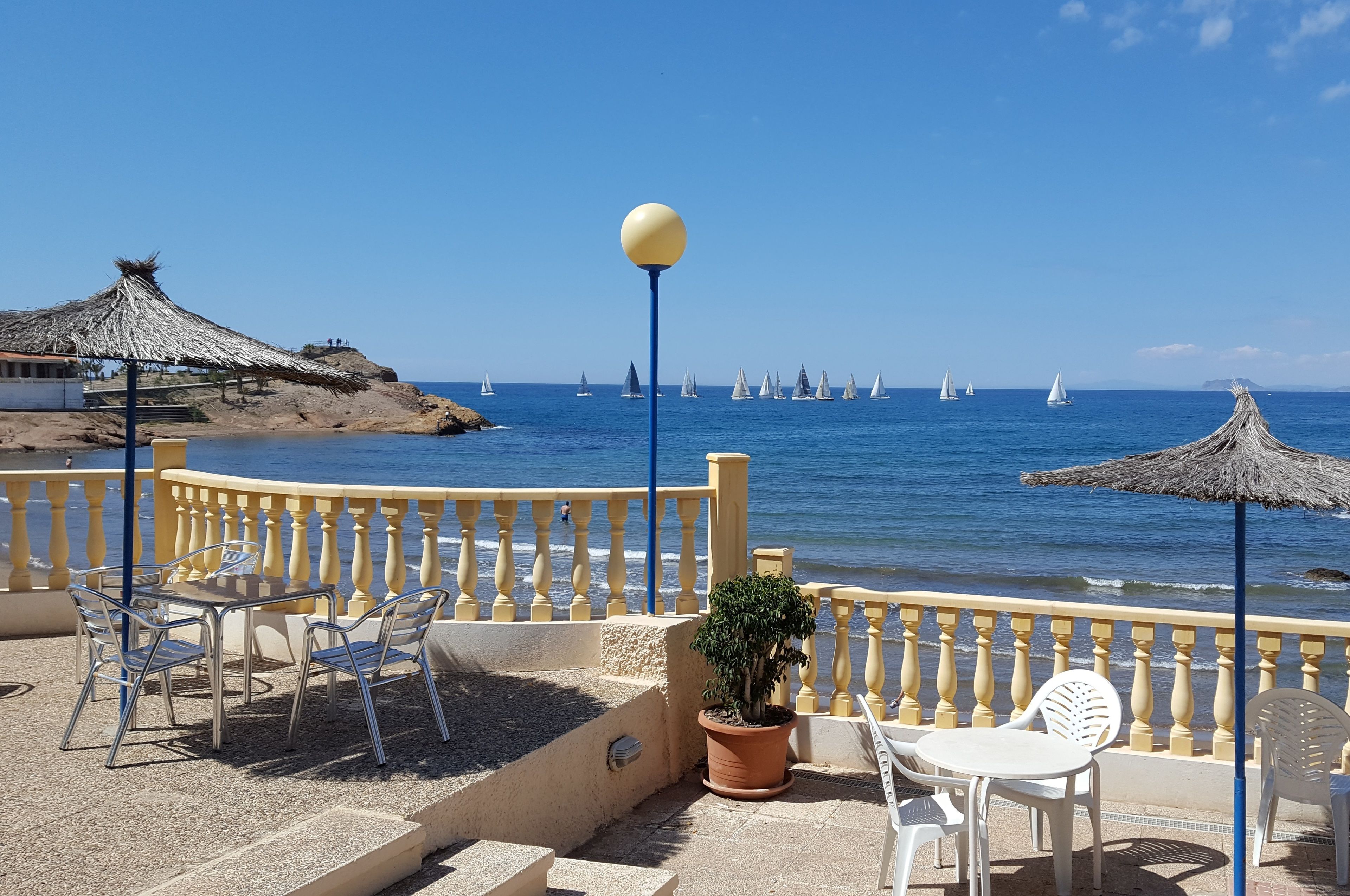 Une terrasse en bord de mer avec une table et des chaises, un pot de fleurs et un parasol en chaume.