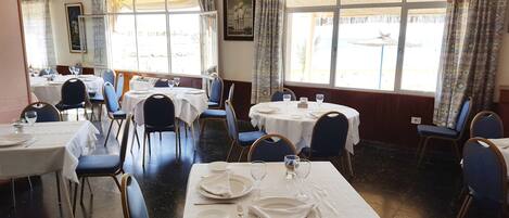 A dining area with round tables and chairs, large windows with curtains, and a framed picture on the wall.