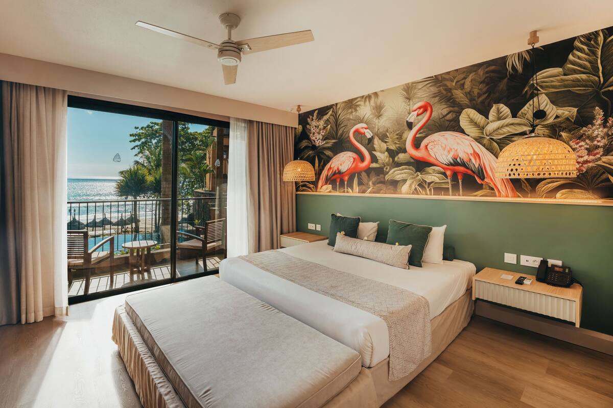 Een hotelkamer met een groot bed, uitzicht op het zwembad en strand, een plafondventilator en een tropische muurschildering.