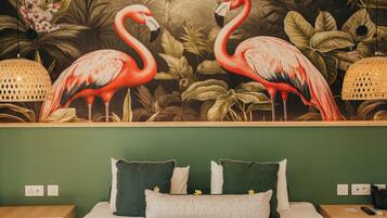 A hotel room with a mural of flamingos, a bed with green and white pillows, and a nightstand with a lamp and phone.