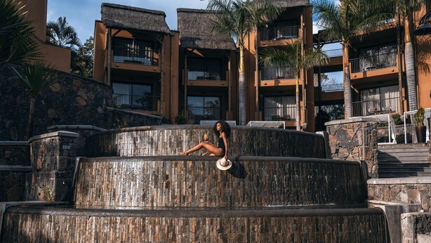 Uma pessoa relaxando em um banco de pedra perto de uma piscina, com um edifício ao fundo.