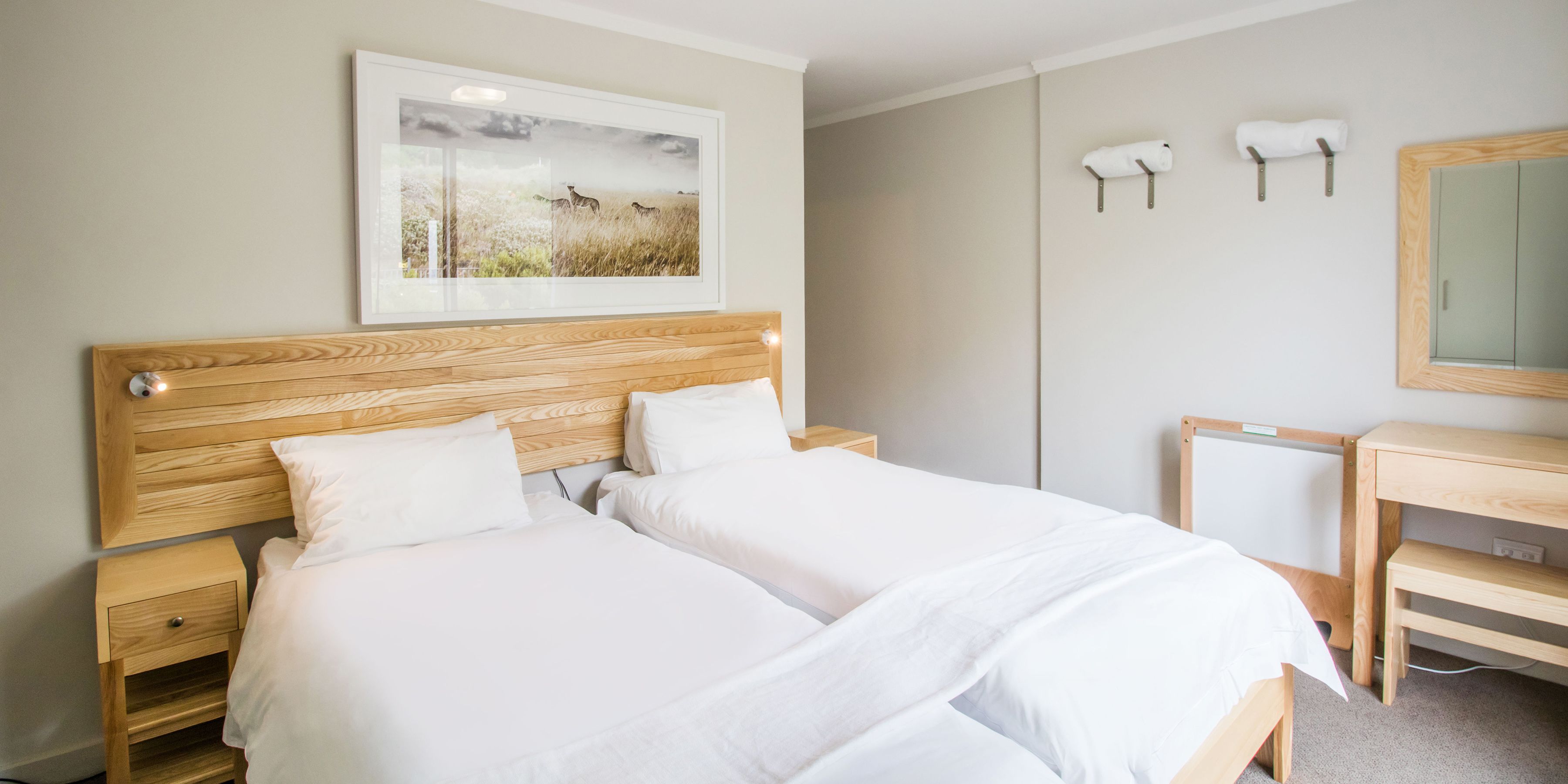 Uma cama de casal com cabeceira de madeira, duas mesas de cabeceira, uma pequena escrivaninha, um espelho e um quadro pendurado na parede.