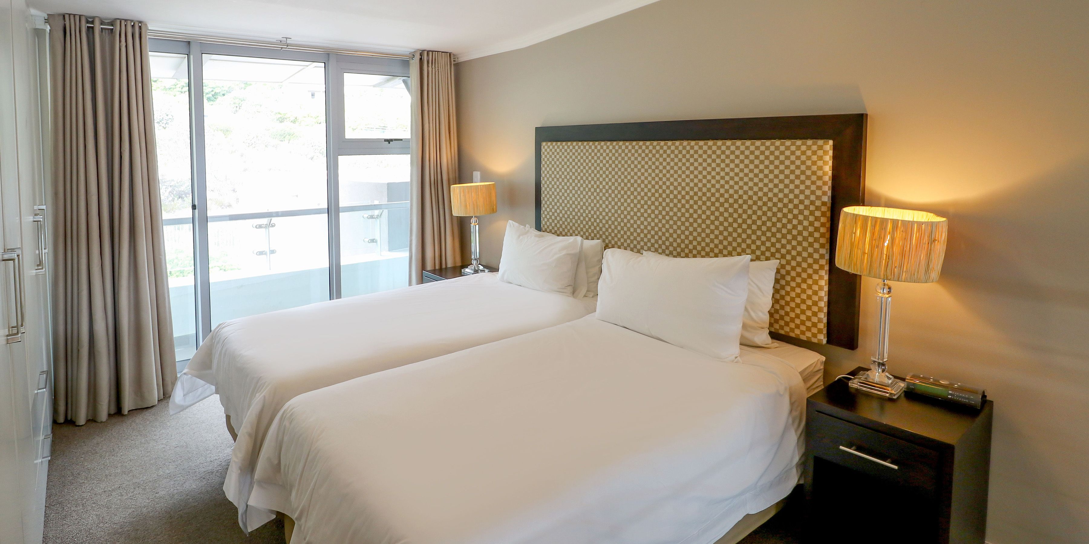 A hotel room with two beds, a large window with curtains, a nightstand with a lamp, and a checkered wall pattern.