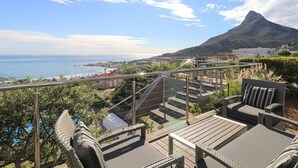 Eine Terrasse mit Gartenmöbeln, einer Glasbrüstung und Blick auf das Meer und ein Gebirge.