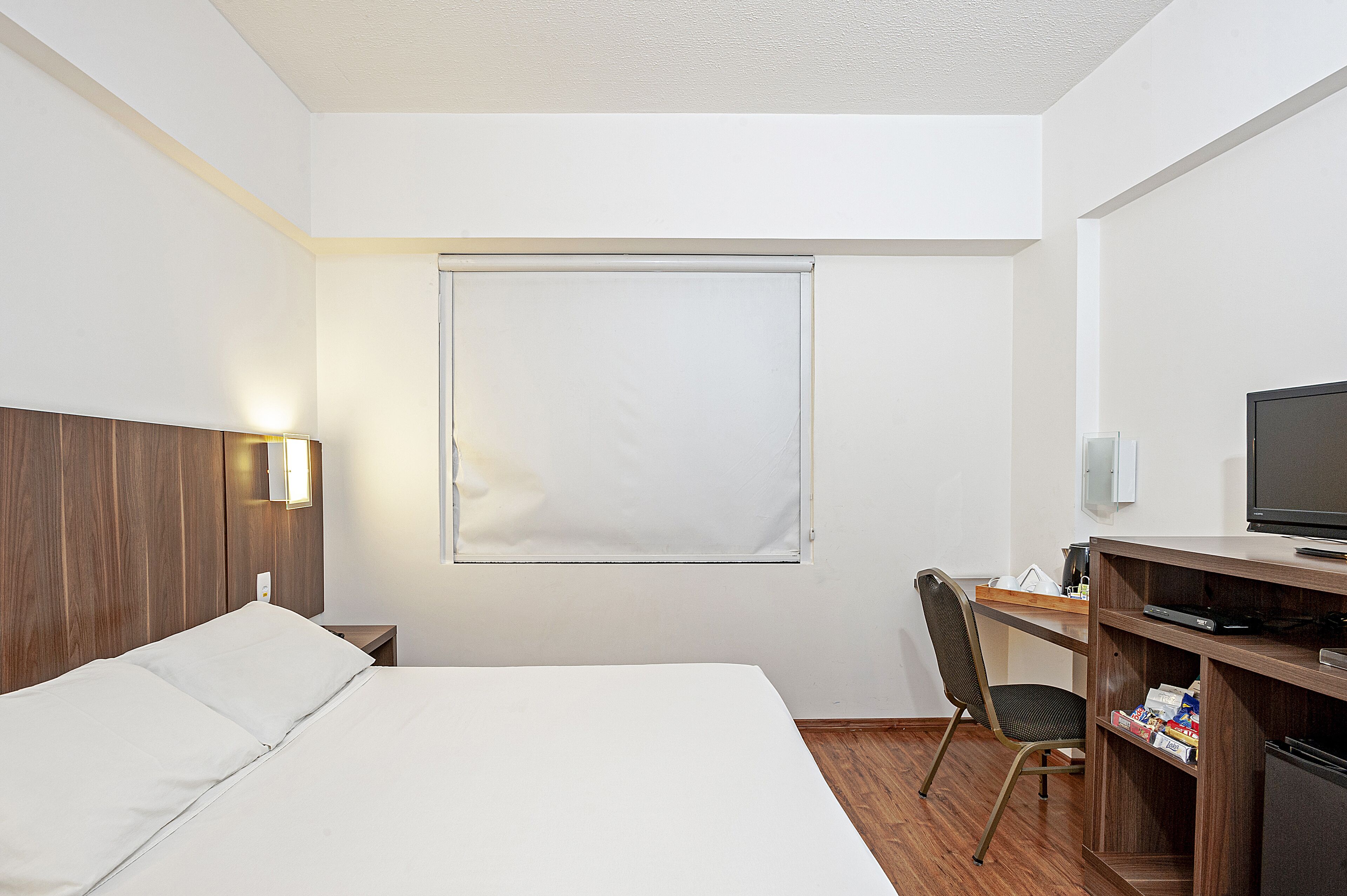 Habitación de hotel con cama, escritorio, silla, televisor y ventana con persianas.