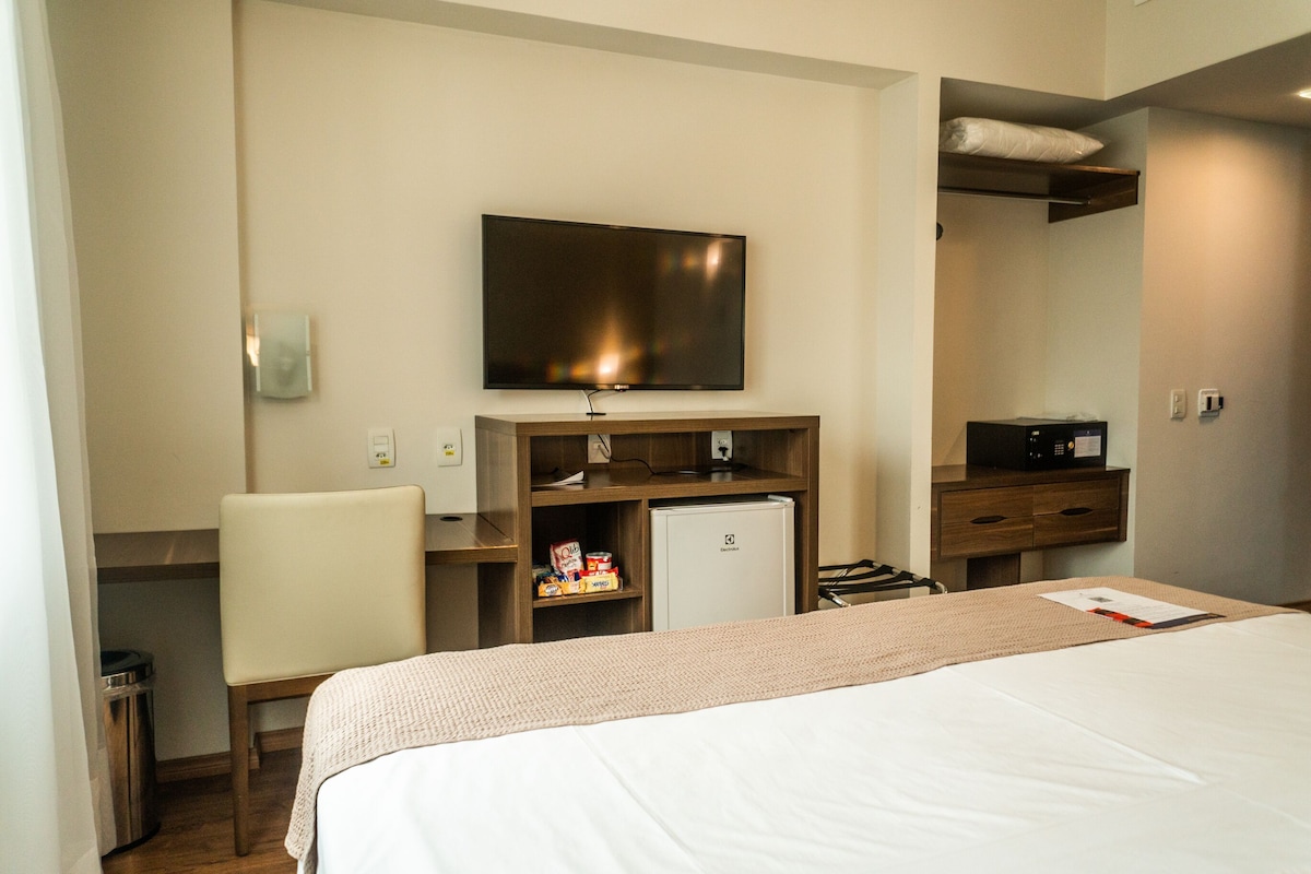 A hotel room with a bed, a television, a small refrigerator, a desk, and a chair.