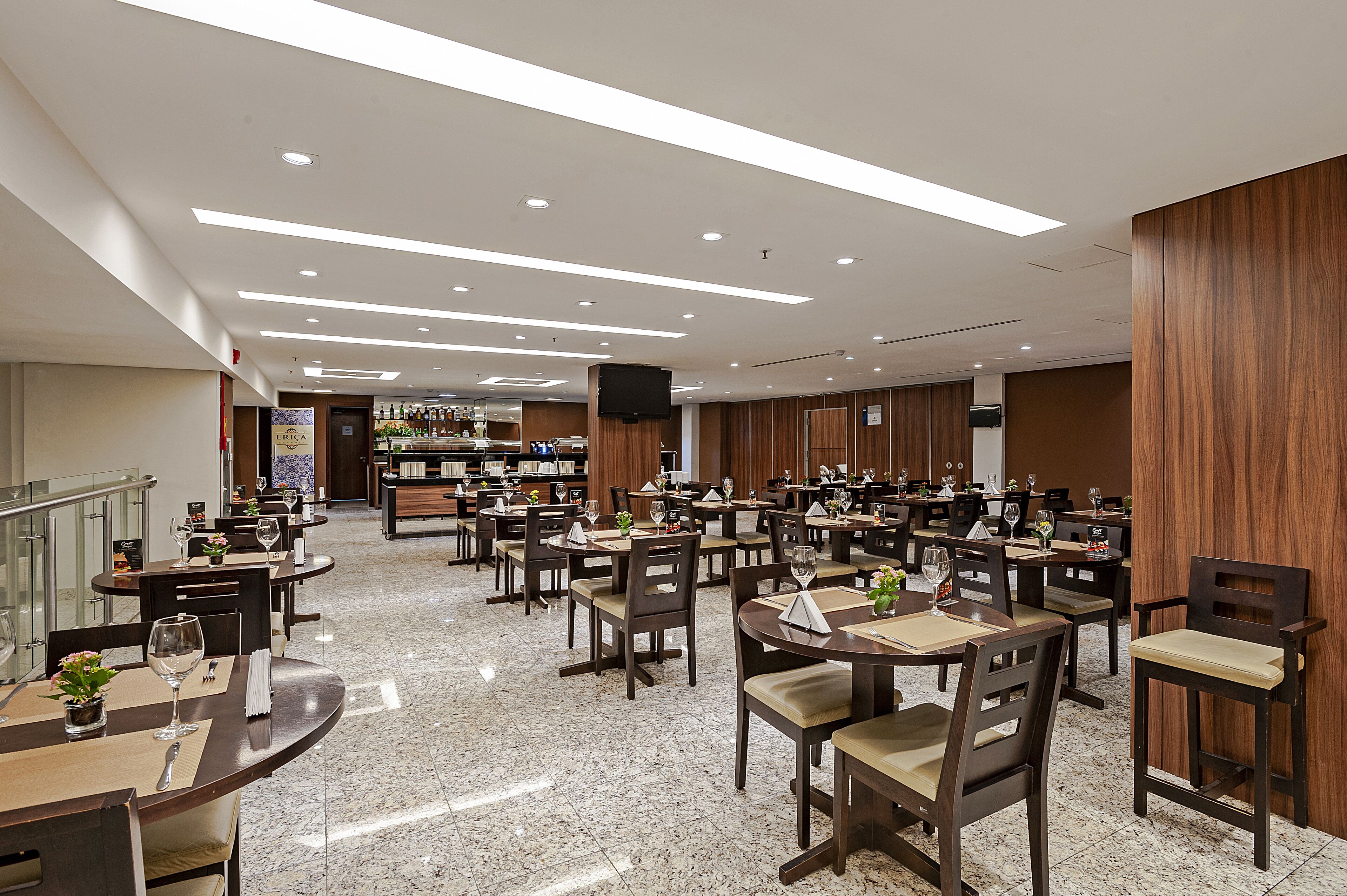 A spacious hotel dining area with multiple tables and chairs, a bar in the background, and a polished marble floor.