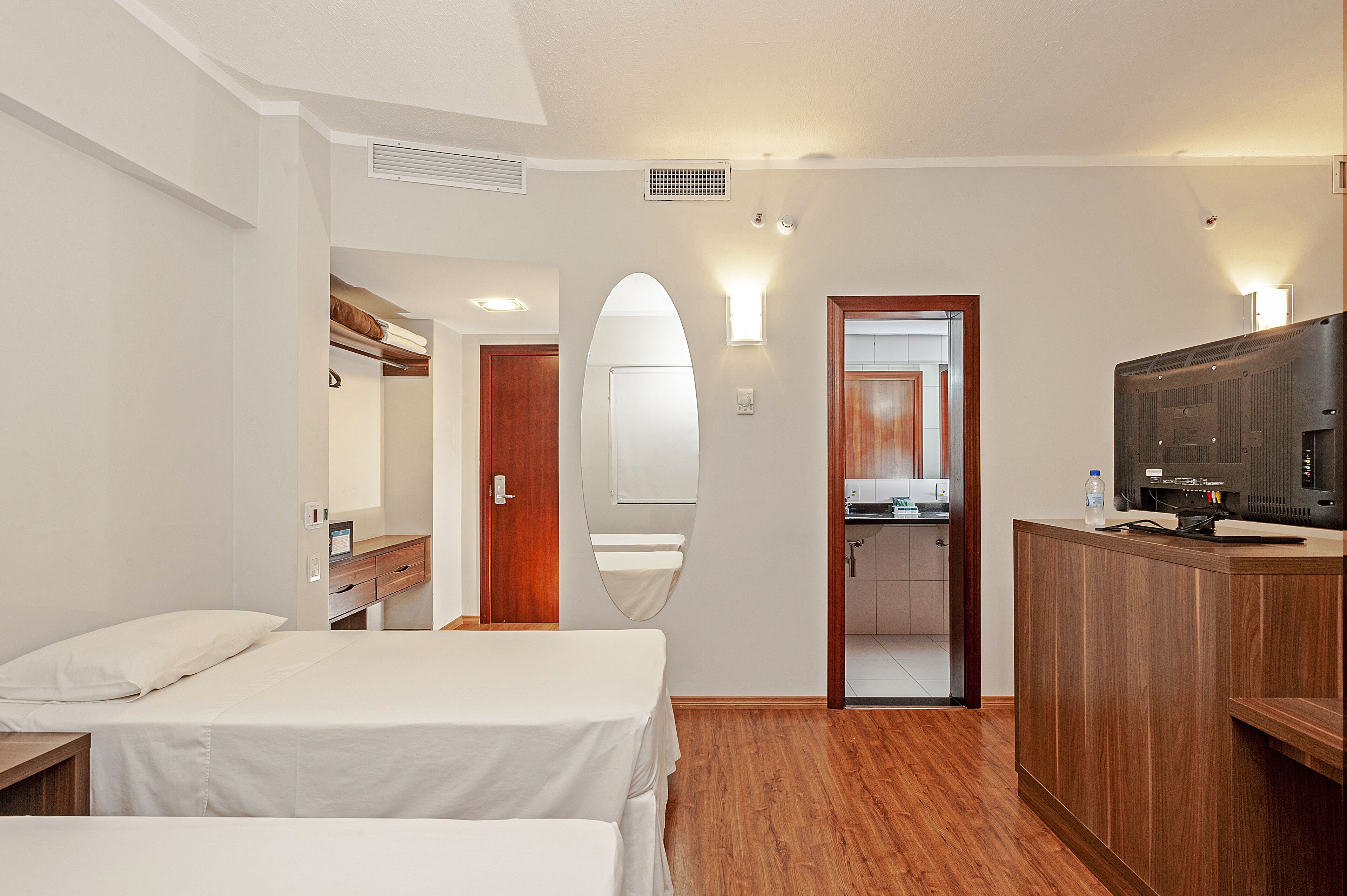 Habitación de hotel con dos camas, un escritorio de madera, un televisor y un baño visible a través de un arco.
