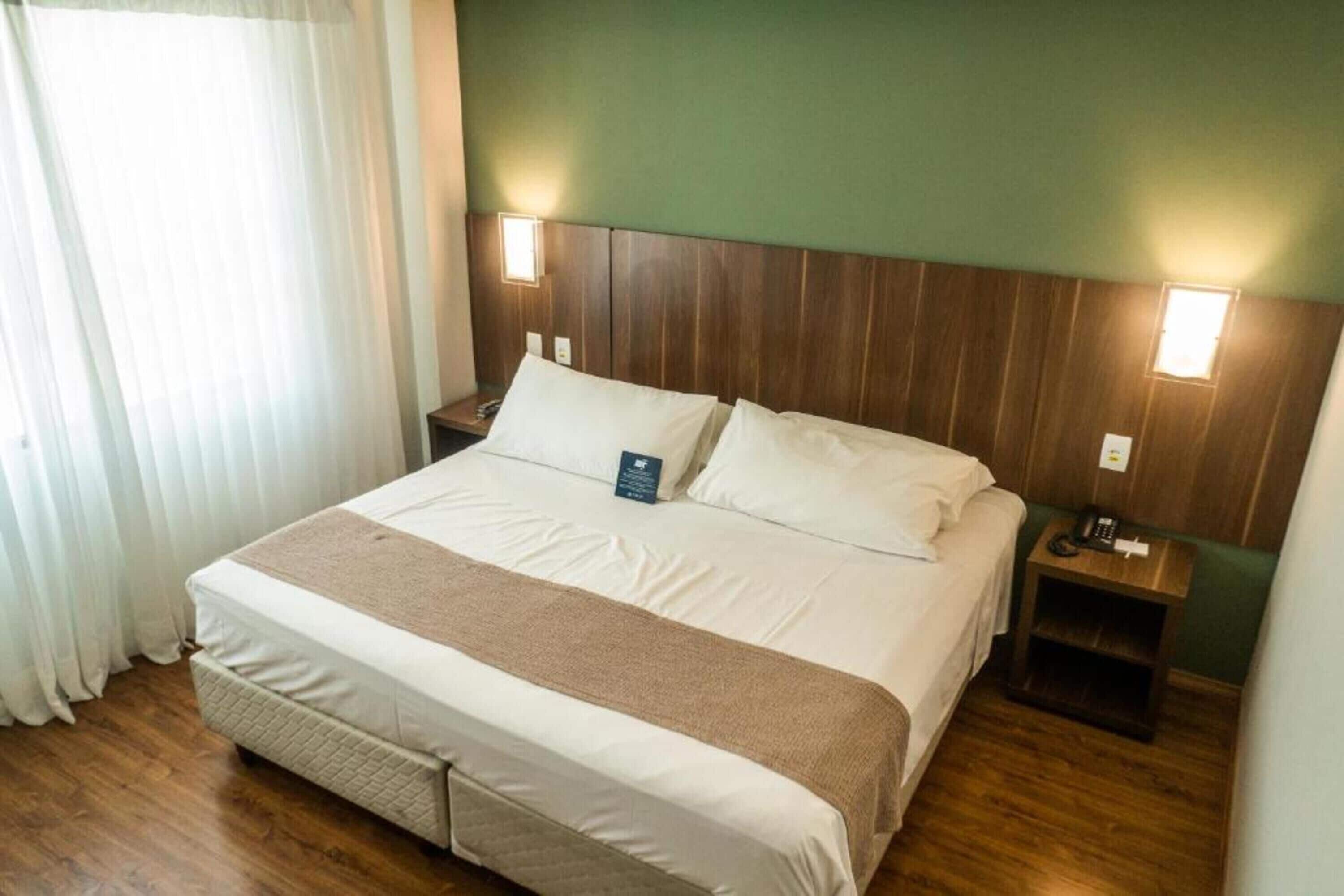A hotel room with a large bed, wooden headboard, bedside tables with lamps, and a window with curtains.