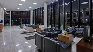 A modern lobby with leather sofas, a reception desk, and large windows offering views of the interior. - Firenze Business Hotel (Palhoça)