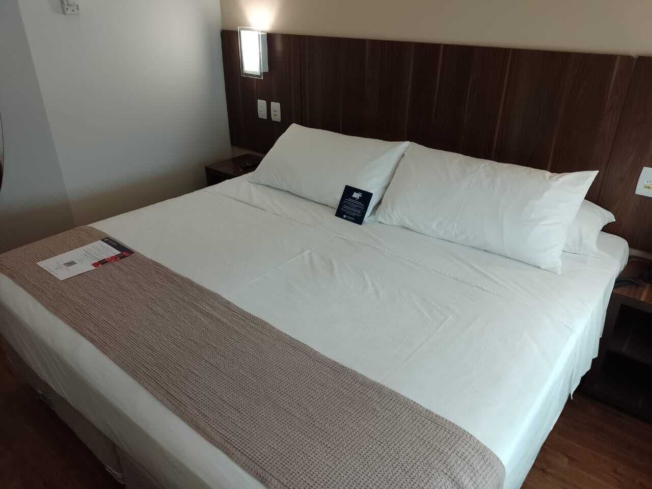 Un letto rifatto con lenzuola bianche e una coperta marrone, una testiera in legno e un comodino con una lampada.
