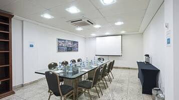 Un salón de conferencias con una larga mesa, sillas, una pantalla de proyector y una mesa negra a la derecha.