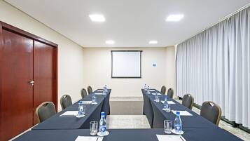 Un salón de conferencias con una larga mesa, sillas, botellas de agua y una pantalla de proyector.